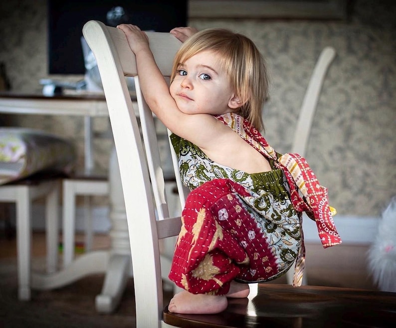 May include: A child wearing a sleeveless romper featuring a patchwork design with red, green, and white floral and geometric patterns. The garment has a tie-back closure and a gathered waist, constructed from quilted fabric with visible stitching.