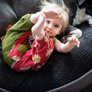 May include: A toddler wearing a long-sleeved romper with a green and red quilted fabric pattern featuring white stitched details. The garment has elasticated cuffs at the wrists and ankles, a round neckline, and a relaxed fit, shown in a casual setting.