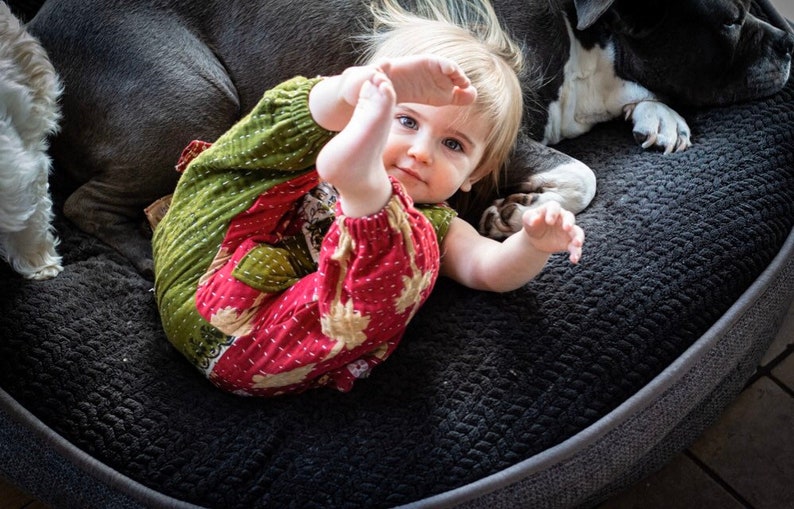 May include: A toddler wearing a long-sleeved romper with a green and red quilted fabric pattern featuring white stitched details. The garment has elasticated cuffs at the wrists and ankles, a round neckline, and a relaxed fit, shown in a casual setting.