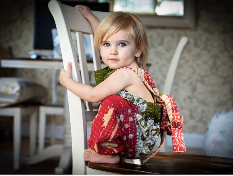 May include: A toddler wearing a sleeveless jumpsuit made of patchwork fabric featuring red, green, and white patterns with visible kantha-style stitching. The garment has a loose, relaxed fit with an elasticised waist and tie-back closure.