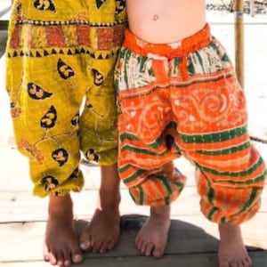 May include: Two pairs of patterned trousers. One is yellow with black leaf designs, the other orange with green stripes and swirls. Both have elasticated waistbands and gathered ankles, suggesting a comfortable fit. The trousers are suitable for children.