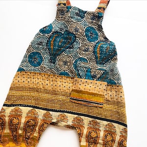 May include: A pair of dungarees featuring a patchwork design with a blue and cream circular floral pattern on the bodice, a mustard yellow diamond-patterned midsection, and a cream-coloured hem with a repeating orange and brown paisley print.