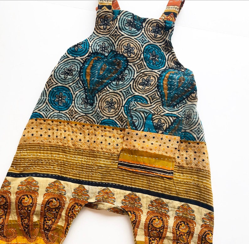 May include: A pair of dungarees featuring a patchwork design with a blue and cream circular floral pattern on the bodice, a mustard yellow diamond-patterned midsection, and a cream-coloured hem with a repeating orange and brown paisley print.