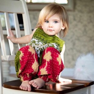 May include: A toddler with blonde hair wearing a sleeveless green top with a white floral pattern and loose-fitting red trousers featuring a gold star-like print. The child is sitting on a white wooden chair with a dark brown seat.