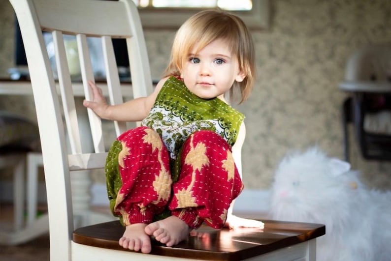 May include: A toddler with blonde hair wearing a sleeveless green top with a white floral pattern and loose-fitting red trousers featuring a gold star-like print. The child is sitting on a white wooden chair with a dark brown seat.