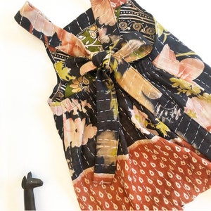 May include: A sleeveless romper featuring a black base with a multi-coloured floral and teardrop pattern. The garment includes a tie-back closure with shoulder straps and white running stitch details throughout the fabric, paired with a small black figurine.