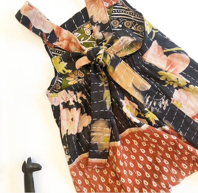 May include: A sleeveless romper featuring a black base with a multi-coloured floral and teardrop pattern. The garment includes a tie-back closure with shoulder straps and white running stitch details throughout the fabric, paired with a small black figurine.
