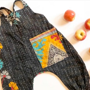 May include: A black sleeveless romper featuring white kantha-style running stitch embroidery throughout. The garment includes a large rectangular patch pocket on the front with a yellow, blue, and black floral and geometric pattern print.