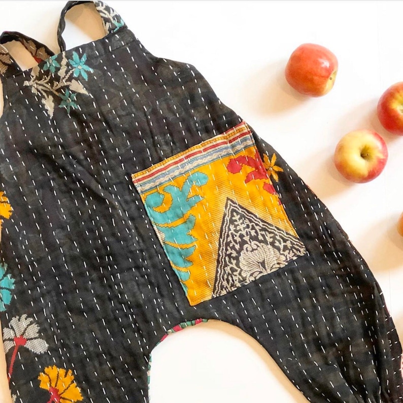 May include: A black sleeveless romper featuring white kantha-style running stitch embroidery throughout. The garment includes a large rectangular patch pocket on the front with a yellow, blue, and black floral and geometric pattern print.