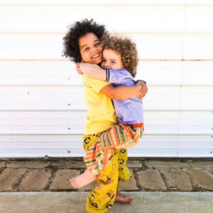 May include: Two young children embrace, one lifting the other. The child on the left wears a yellow shirt and patterned pants. The child being held wears a blue shirt and patterned pants. Both children are smiling.