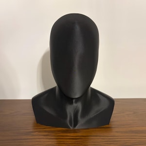 May include: A black, faceless bust, likely for display or storage of wigs, hats, or other headwear. The bust is made of a smooth, solid material and sits on a wooden surface. The bust has a neck and shoulders.