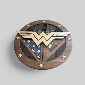 May include: A round wooden display case with a glass front, featuring a Wonder Woman logo and star accents. Inside, it holds a folded American flag and a military emblem. Text includes "Director, HR 01.07.2008" and "Sandra D. Brown 12.31.2025".