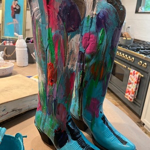 May include: A pair of blue cowboy boots with colorful abstract paint designs. The boots have a pointed toe and a high heel.