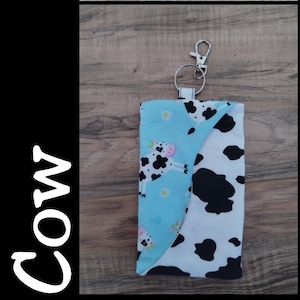 May include: A blue and white cow print fabric key chain with a silver key ring. The fabric is a light blue with white daisies and black and white cows. The other side of the key chain is a white fabric with black cow spots.