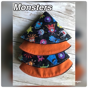 Puede incluir: Tres agarraderas de olla de tela naranja y negra con un estampado de monstruos. Las agarraderas de olla están apiladas una encima de la otra. La agarradera de olla superior tiene un fondo negro con monstruos coloridos sobre ella. Las dos agarraderas de olla inferiores son de color naranja sólido.