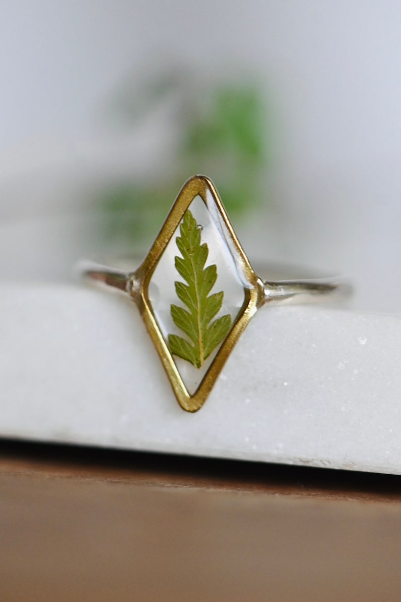 K&ouml;nnte beinhalten: Ein goldener Ring mit einer klaren, rautenf&ouml;rmigen Fassung, in der ein gepresstes gr&uuml;nes Blatt zu sehen ist.