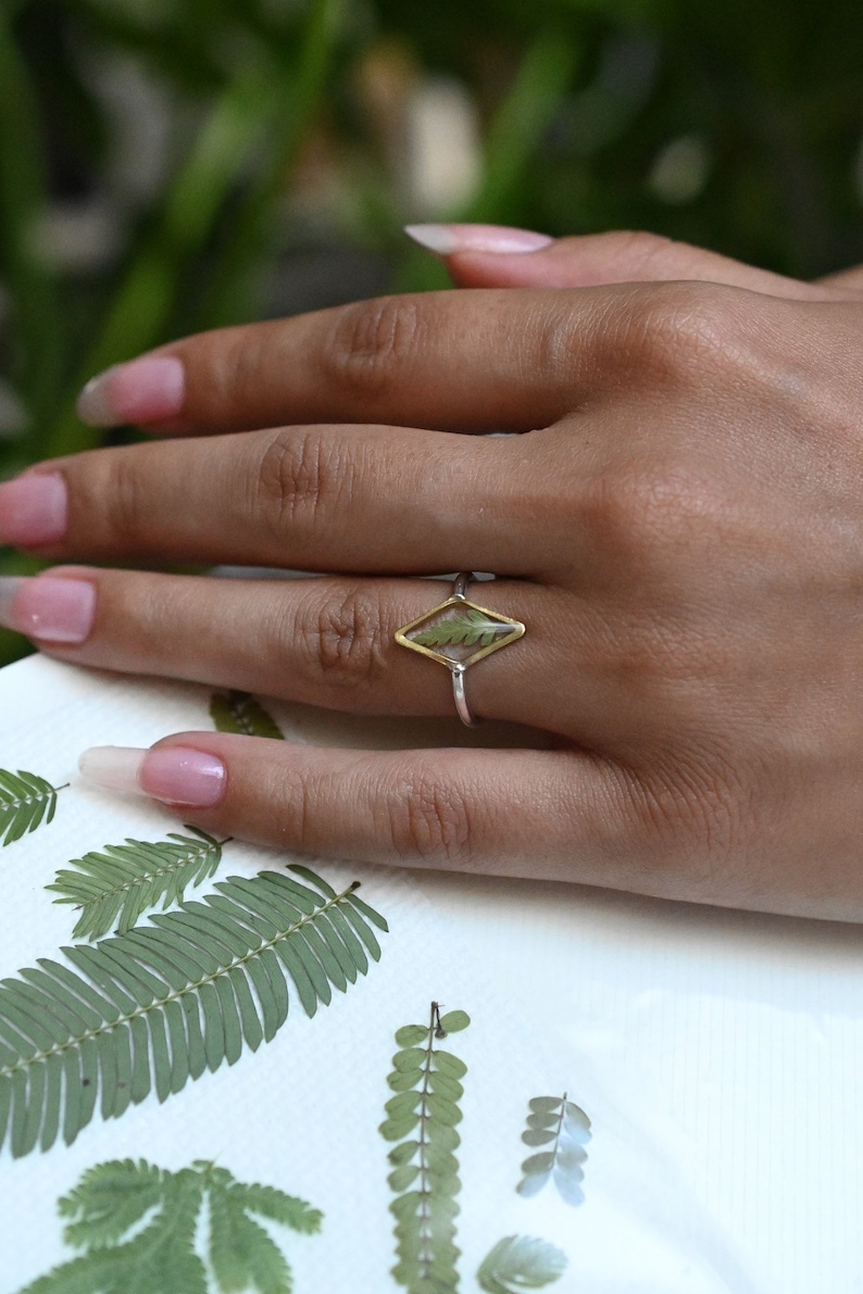 K&ouml;nnte beinhalten: Ein silberner Ring mit einer goldenen, rautenf&ouml;rmigen Fassung, in der ein gepresstes gr&uuml;nes Blatt eingefasst ist. Der Ring befindet sich an einer Hand mit manik&uuml;rten N&auml;geln.