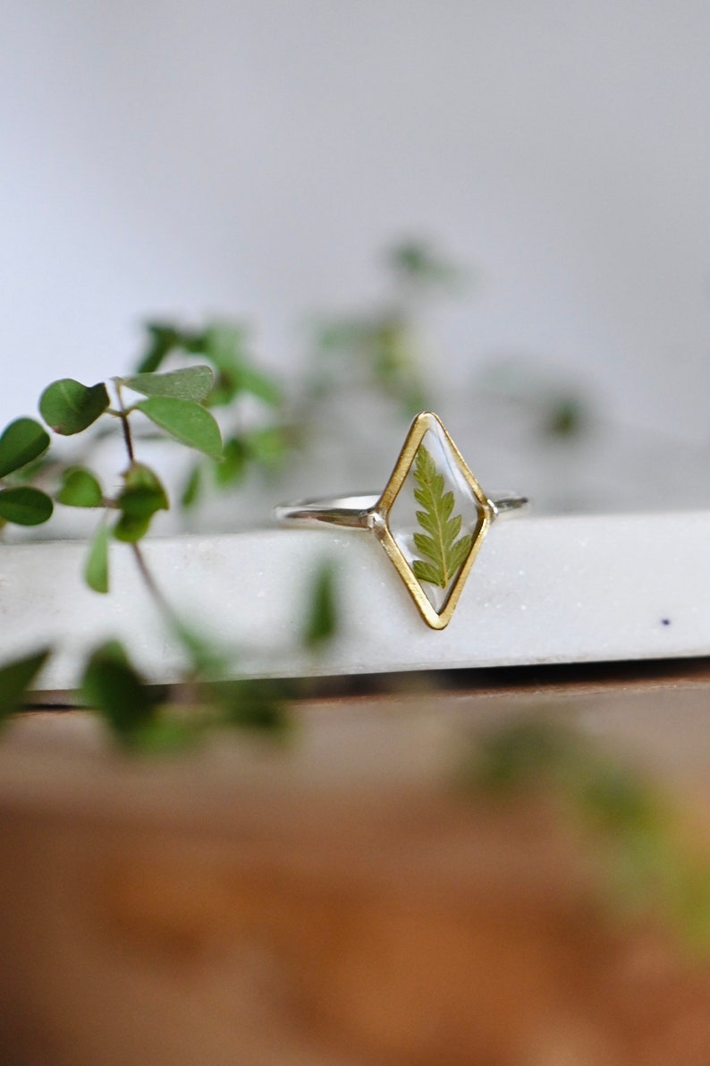 K&ouml;nnte beinhalten: Ein goldener Ring mit einer rautenf&ouml;rmigen Fassung, in der eine gepresste gr&uuml;ne Farnpflanze eingearbeitet ist. Der Ring liegt auf einer wei&szlig;en Oberfl&auml;che.