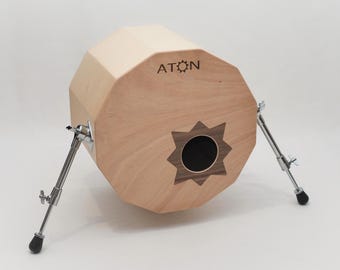 Op de afbeelding: Een achtehoekige houten trommel met een zwart gat in het midden en het woord "ATON" erop gegraveerd. De trommel wordt ondersteund door twee metalen poten.