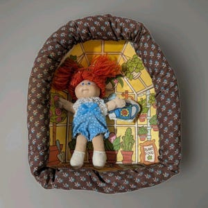 May include: A doll with orange yarn hair and a blue polka-dot romper with white lace trim, resting inside a brown fabric-covered cradle featuring a small blue floral pattern. The background shows a yellow greenhouse illustration with plants.