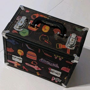 May include: A rectangular black vanity case featuring a repeating pattern of colorful handbags, shoes, and accessories. The case has a black top handle, silver-toned metal corner reinforcements, and two front latch closures with a small silver logo plate.