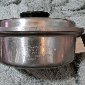 May include: A stainless steel saucepan with a black handle and lid knob. The side is engraved with Duncan Hines STAINLESS STEEL COOKWARE (3-PLY 18-8) REGAL WARE INC. LICENSEE KEWASKUM, WIS. The metal surface has a polished finish.