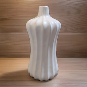 May include: A white ceramic vase with a smooth, matte finish and a distinct vertical ribbed or fluted texture. The vessel features a narrow neck that gradually widens toward the base, creating a symmetrical, elongated, and rounded silhouette.