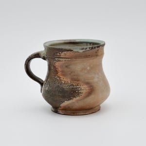 May include: A handmade ceramic mug with a rounded body and a handle. The mug features a blend of brown, tan, and dark gray colors, with a slightly textured surface. The rim has a hint of green. A unique piece for enjoying beverages.