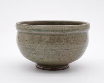 May include: A handmade ceramic bowl with a rounded shape and a small, circular base. The bowl has a muted green and brown color palette, with a slightly textured surface. The rim of the bowl is a darker shade of green.