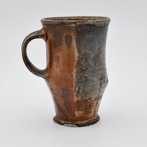 May include: A handmade ceramic mug with a unique, asymmetrical design. The mug features a brown and gray color scheme, with a textured surface and a sturdy handle. The mug is approximately 5 inches tall.