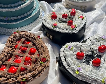 May include: Five heart-shaped purses decorated to look like different types of cakes. The purses are on a white tablecloth. One purse is a chocolate cake, one is a white cake with cherries, one is a cherry pie, one is a black cake with white frosting and cherries, and one is a blue cake with white frosting.