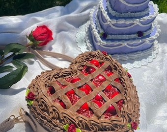 May include: A heart-shaped cherry pie purse with a brown chocolate crust and red cherries. The purse has a brown strap and is decorated with small red roses.