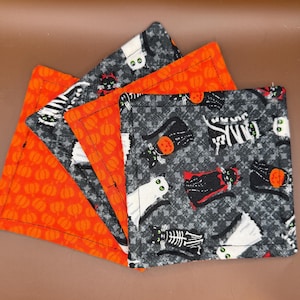 Könnte beinhalten: Set aus vier Halloween-Servietten. Zwei Servietten zeigen orangefarbene Kürbismuster, während die anderen beiden Designs mit schwarzen Katzen und Skeletten auf grauem Hintergrund aufweisen. Jede Serviette hat einen schwarz genähten Rand.