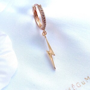 May include: Gold hoop earrings with a dangling lightning bolt charm. The hoop is encrusted with clear cubic zirconia stones.
