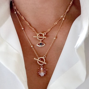 May include: A layered gold-tone necklace set featuring two chains with toggle clasps. The top chain includes a small evil eye pendant with a dark blue center and clear stones. The bottom chain features a spider pendant with a clear stone center.