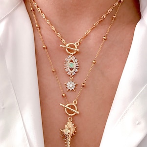 May include: A layered gold-tone necklace set featuring two chains. The top chain has a toggle clasp with an eye-shaped pendant containing clear crystals and a central light green stone, plus a star-shaped charm. The bottom chain features a seashell pendant.