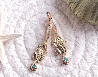 May include: Gold hoop earrings with a seahorse charm and a blue gemstone. The earrings are on a white textured surface.