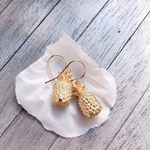 Könnte beinhalten: Goldfarbene Ohrringe in Form von Ananas mit strukturierter Oberfläche, auf einem weißen Tuch liegend. Die Ohrringe haben einen Hakenverschluss und sind vor einem hellgrauen Holzgrund platziert. Tropischer Schmuck.