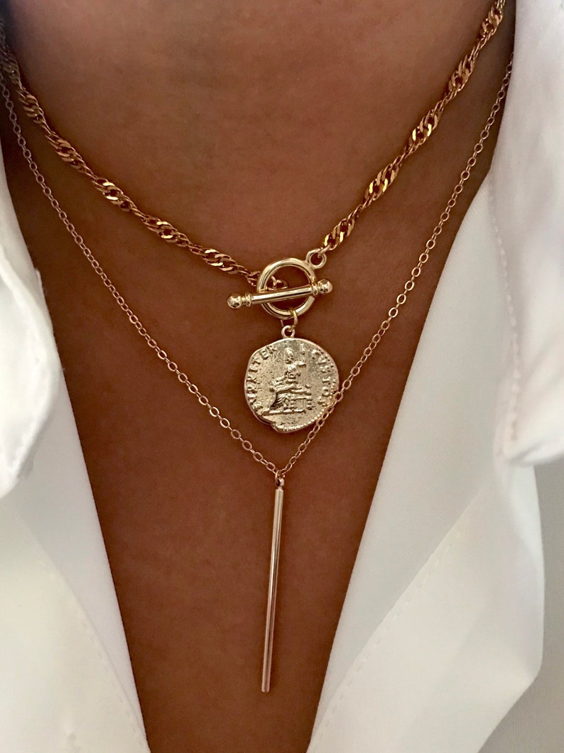 May include: A layered gold-tone necklace set featuring two distinct chains. The top chain is a twisted rope style with a toggle clasp and a circular coin pendant. The bottom chain is a delicate link style with a smooth, vertical bar pendant.