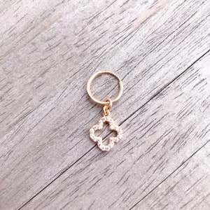 May include: Gold hoop earring with a dangling charm in the shape of a four-leaf clover. The clover is encrusted with clear rhinestones.