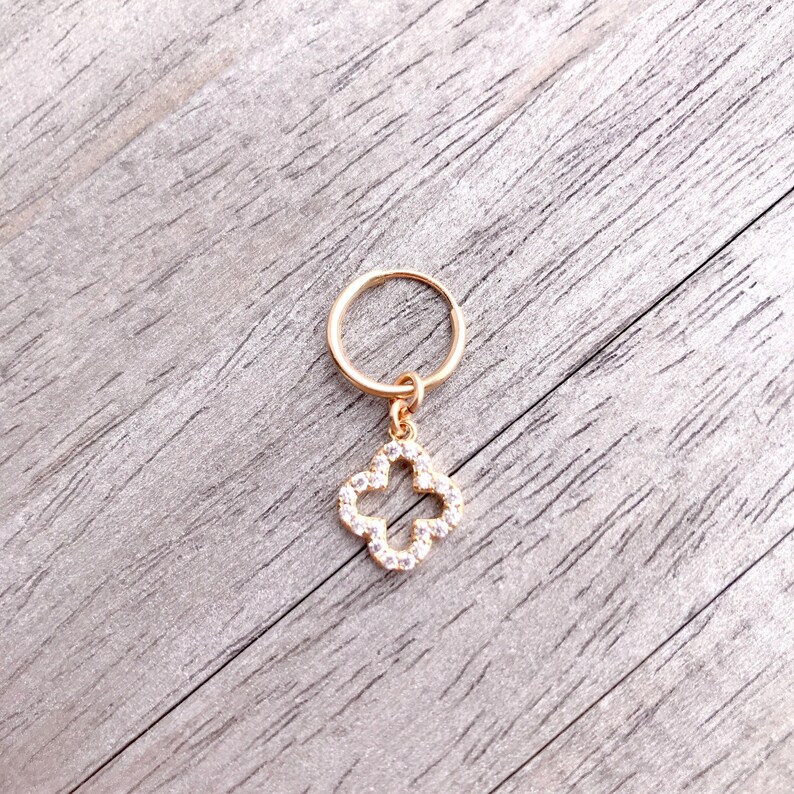 May include: Gold hoop earring with a dangling charm in the shape of a four-leaf clover. The clover is encrusted with clear rhinestones.