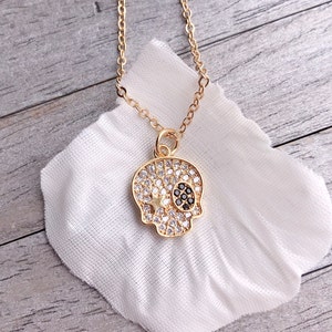 Op de afbeelding: Een goudkleurige ketting met een schedel hanger. De schedel is bezet met heldere en zwarte edelstenen, met een ster accent. De ketting is een sieraad en ligt op een witte doek.