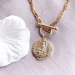 May include: A gold-toned necklace featuring a twisted chain and a toggle clasp. A circular pendant hangs from the toggle, embossed with a classical figure seated on a throne and Latin-style lettering around the edge of the coin-shaped charm.