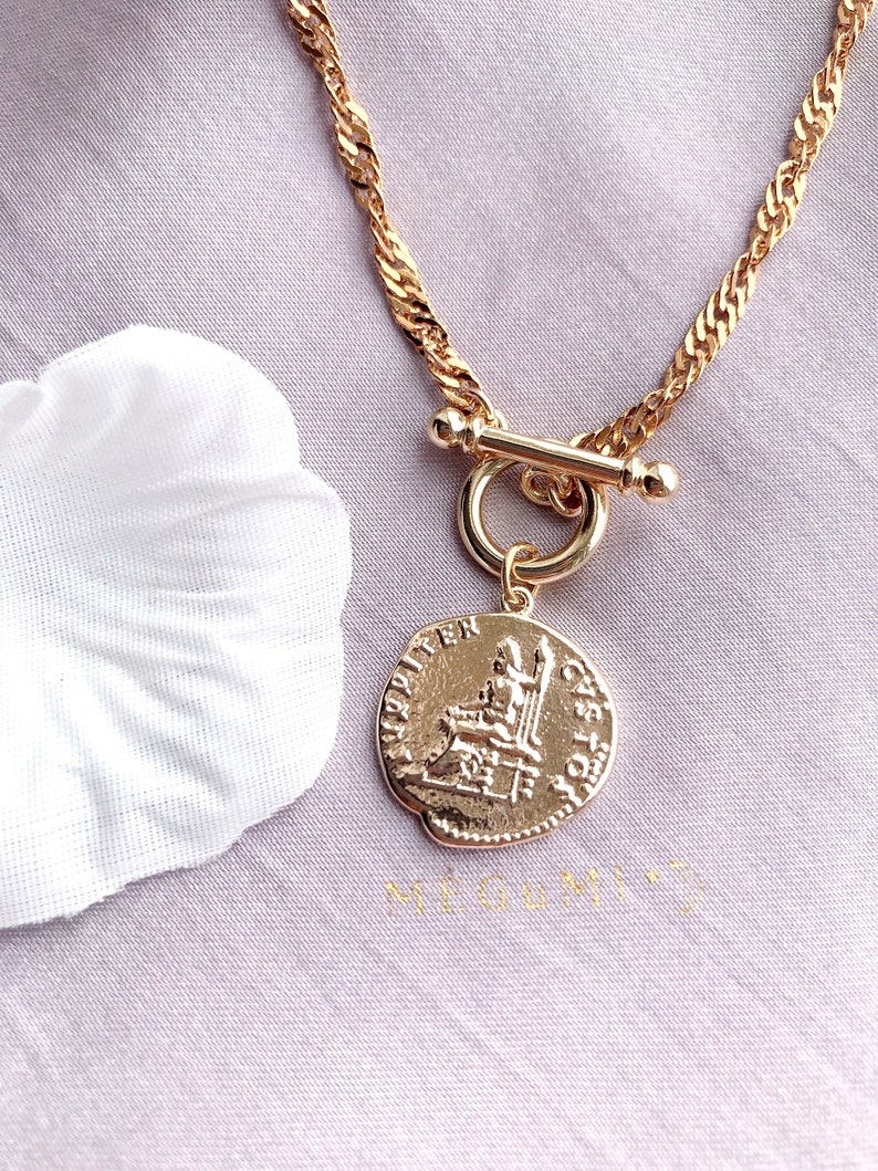 May include: A gold-toned necklace featuring a twisted chain and a toggle clasp. A circular pendant hangs from the toggle, embossed with a classical figure seated on a throne and Latin-style lettering around the edge of the coin-shaped charm.