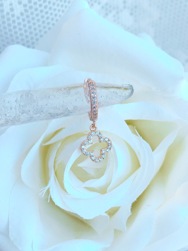 Puede incluir: Aretes de aro de oro rosa con un dije de tr&eacute;bol de cuatro hojas colgante, ambos adornados con piedras de circonita c&uacute;bica transparentes.