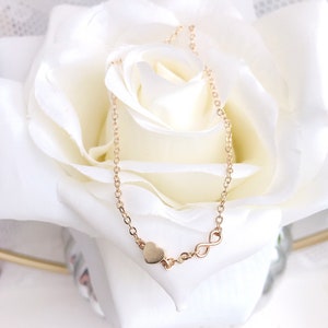 May include: A gold chain necklace with a heart and infinity symbol charm. The necklace is on a white rose.