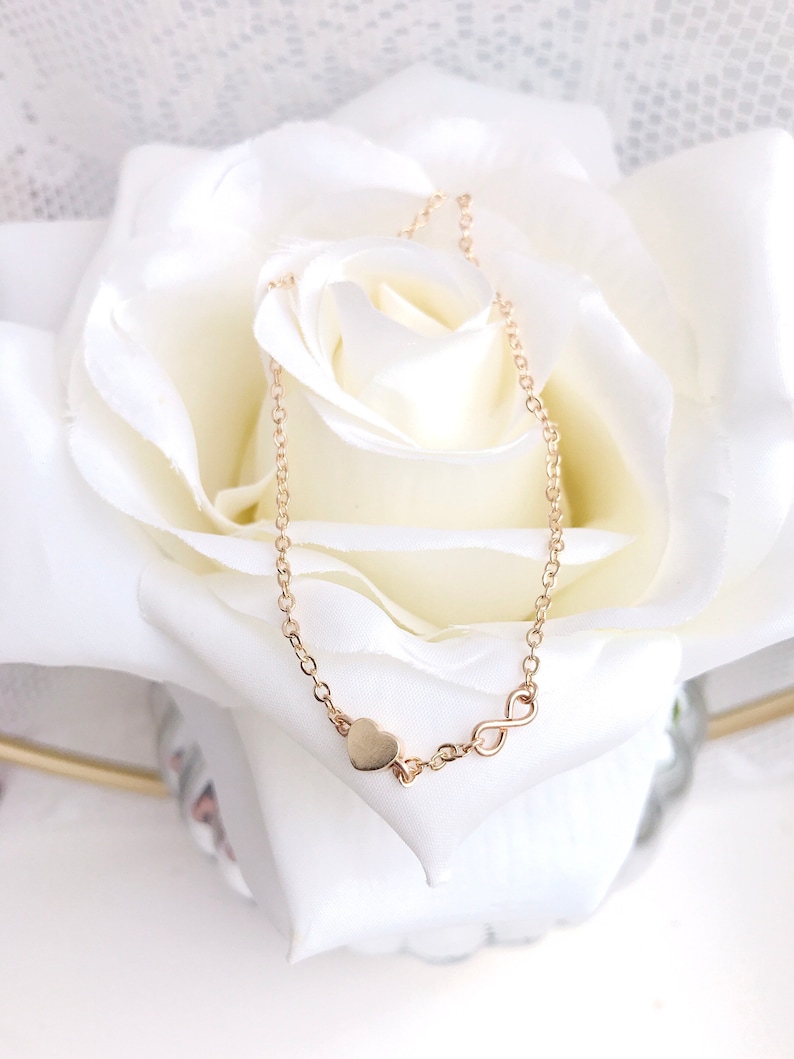 May include: A gold chain necklace with a heart and infinity symbol charm. The necklace is on a white rose.