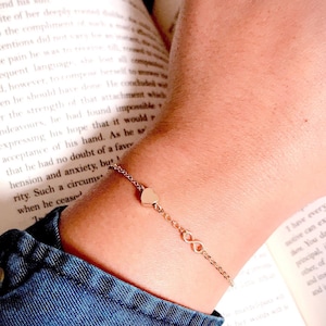 May include: A gold chain bracelet with a heart charm and an infinity symbol charm. The bracelet is on a person's wrist.