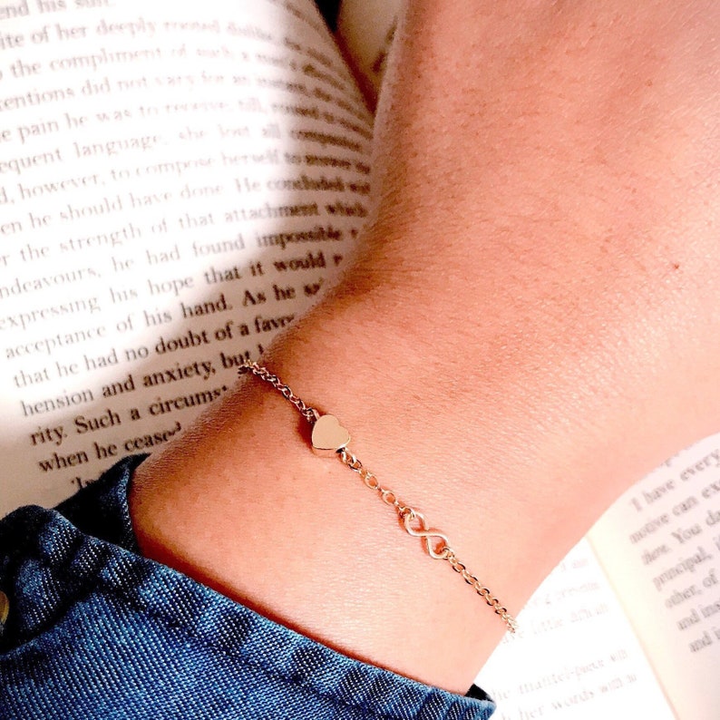 May include: A gold chain bracelet with a heart charm and an infinity symbol charm. The bracelet is on a person's wrist.