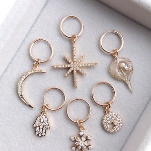 May include: Six gold hoop earrings with different charms. The charms include a crescent moon, a star, a seashell, a hamsa hand, a snowflake, and a circle.